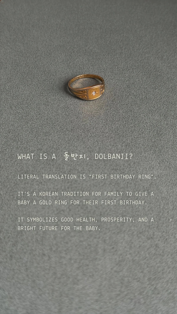 Gold ring on a gray background with text about Korean tradition of giving a gold ring for a baby's first birthday.